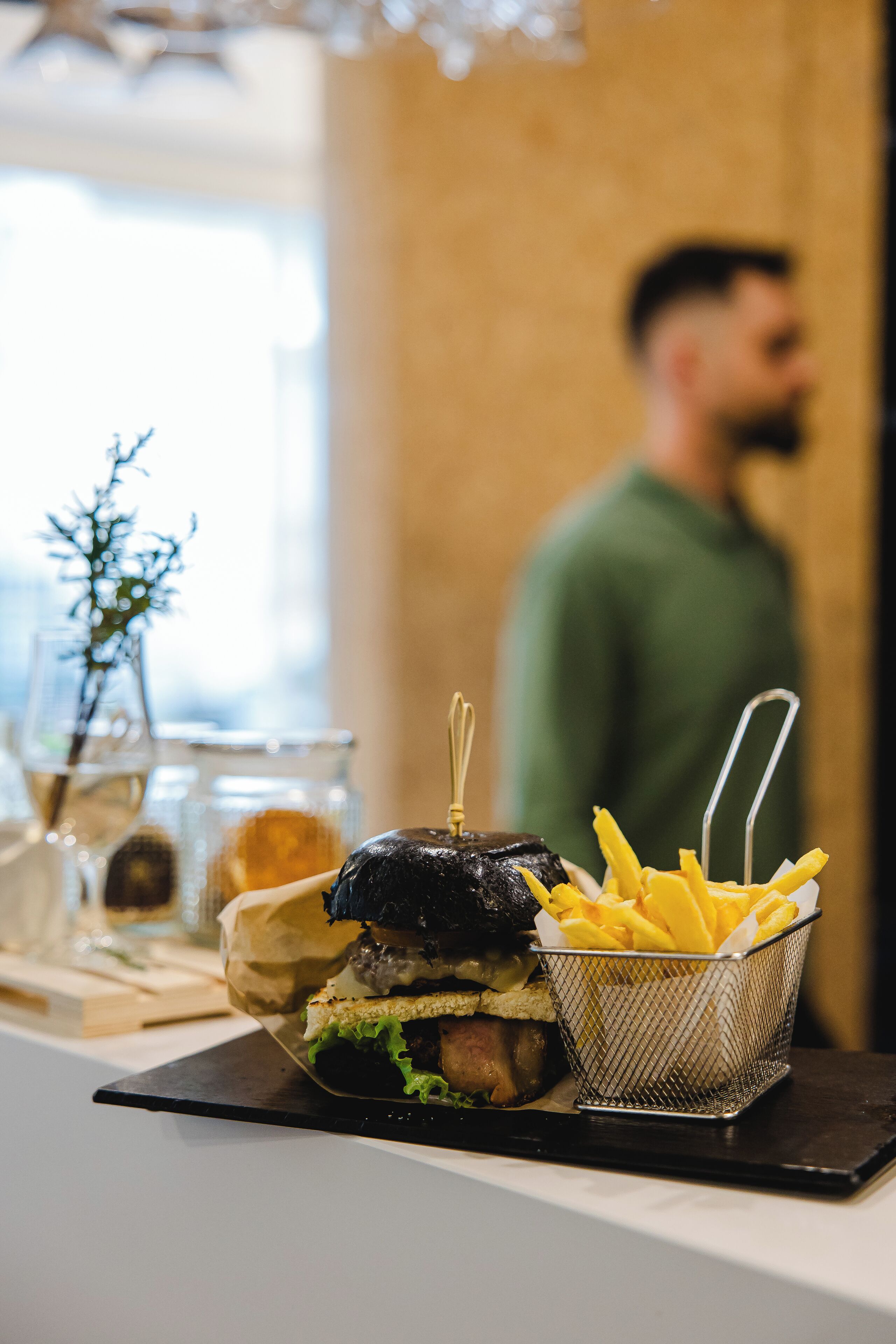 A burger with fries on a black plate, a glass of water, and a vase with a plant in the background.