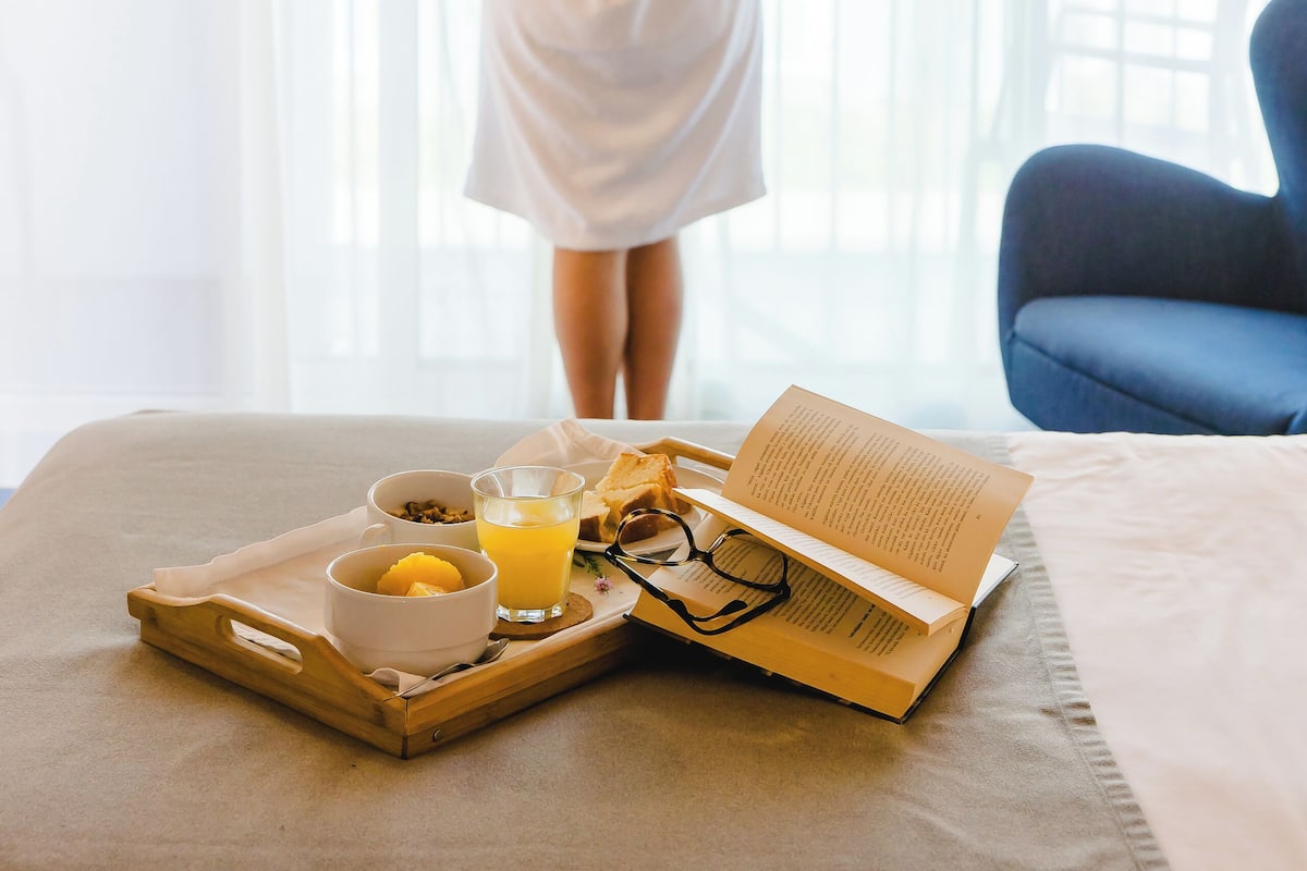 A person in a white robe standing by a window, holding curtains. A breakfast tray with a book, glasses, and orange juice is on the bed.