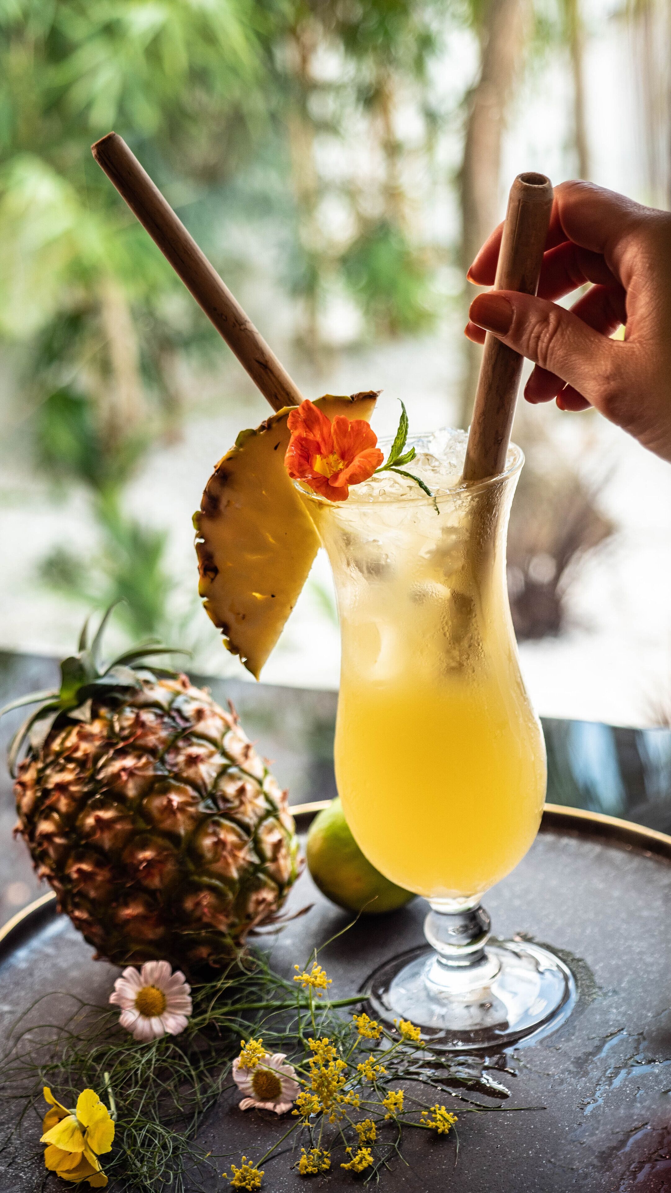 A tropical drink in a glass with a pineapple slice and a flower garnish, a hand holding a straw, and a pineapple on the table.