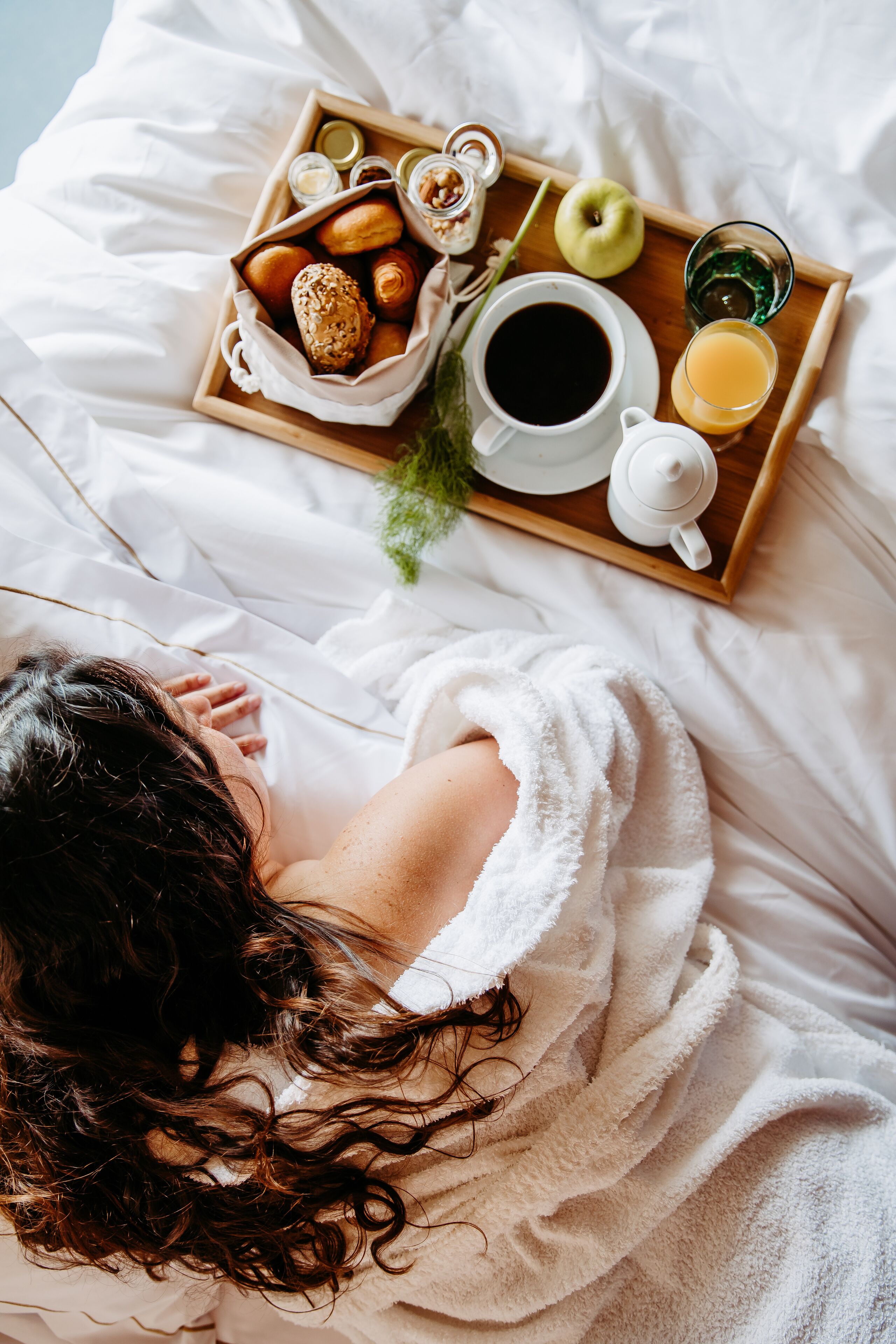 En person ligger i sengen med et bakkehåndtag med morgenmadsprodukter, blandt andet kager, kaffe og juice.