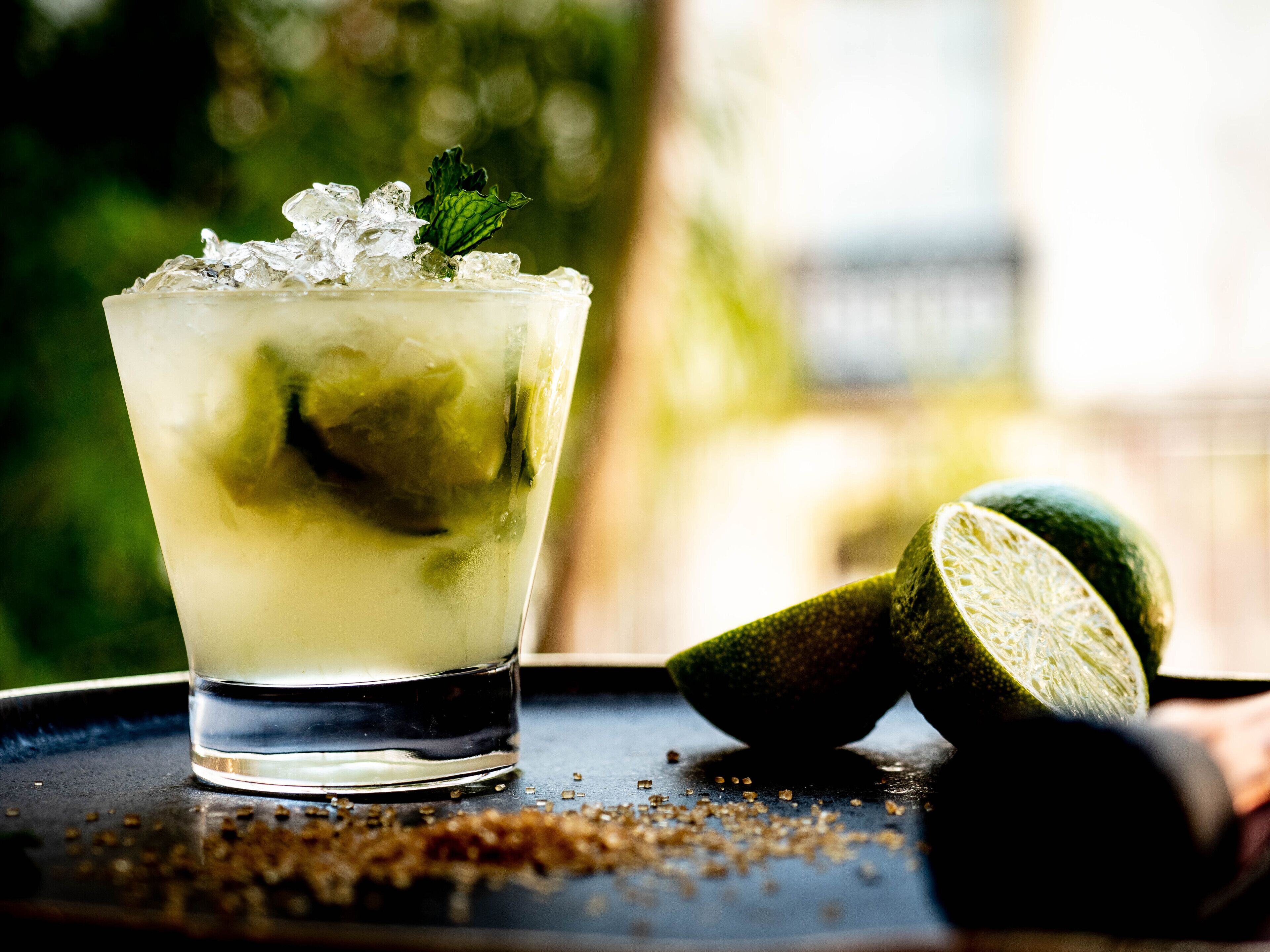 A glass of a lime-based drink with ice cubes and a mint leaf, accompanied by whole and halved limes on a dark surface.