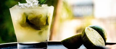 A glass of a lime-based drink with ice cubes and a mint leaf, accompanied by whole and halved limes on a dark surface.