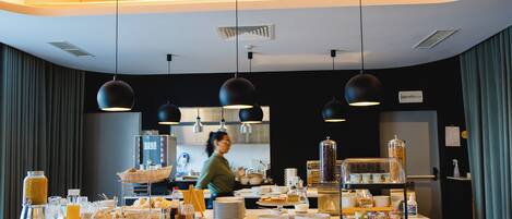 Un bufé de desayuno con diversas opciones de comida y bebidas, un barista preparando bebidas y una cocina moderna al fondo.