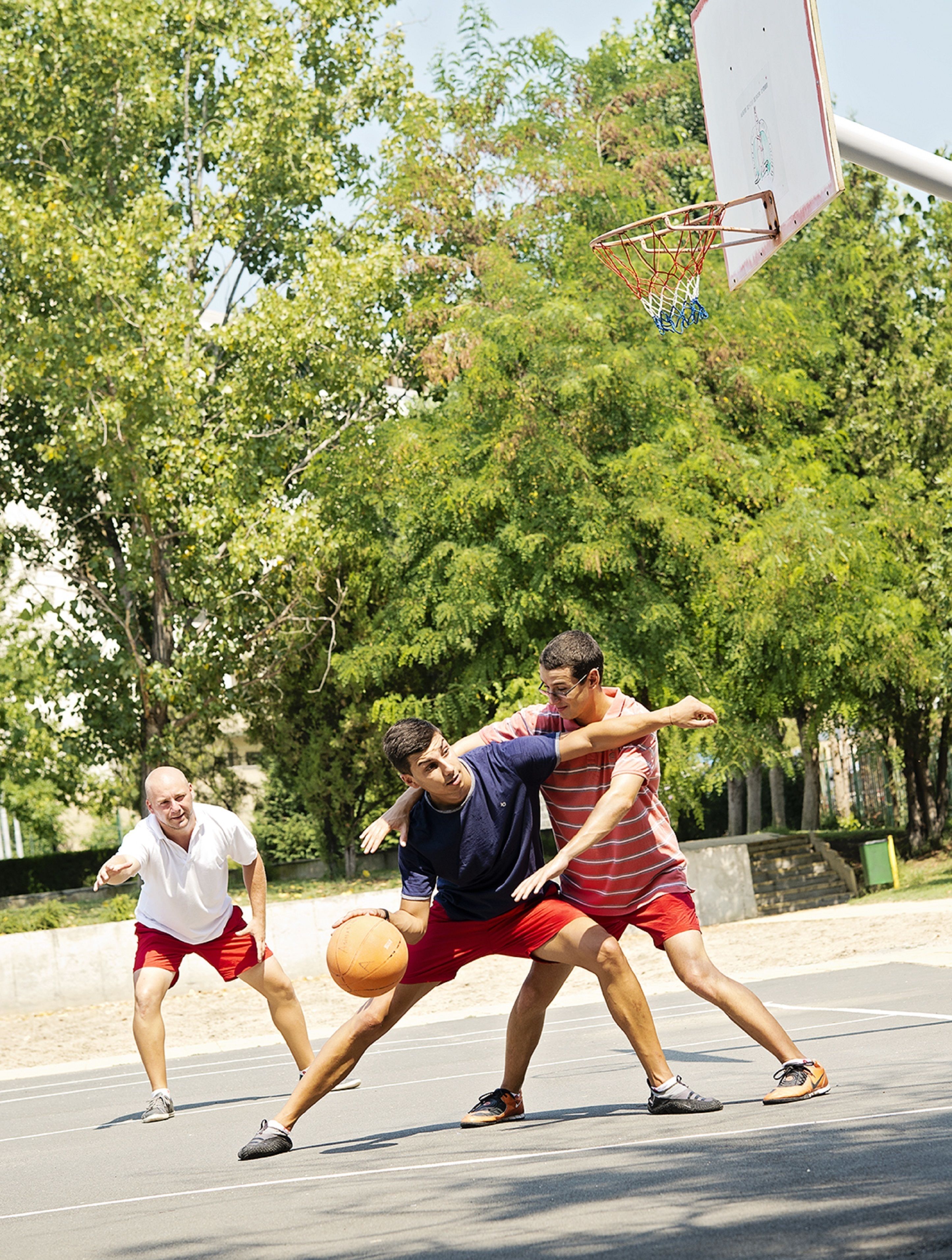 A basketball court with three players in action, trees in the background, and a basketball hoop.