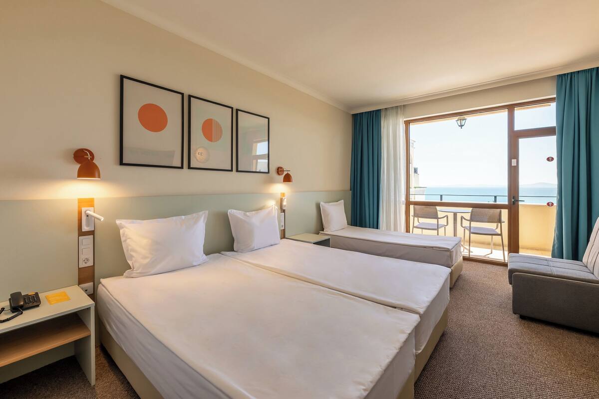 A hotel room with two beds, a sofa, a balcony with a table and chairs, and framed artwork on the wall.