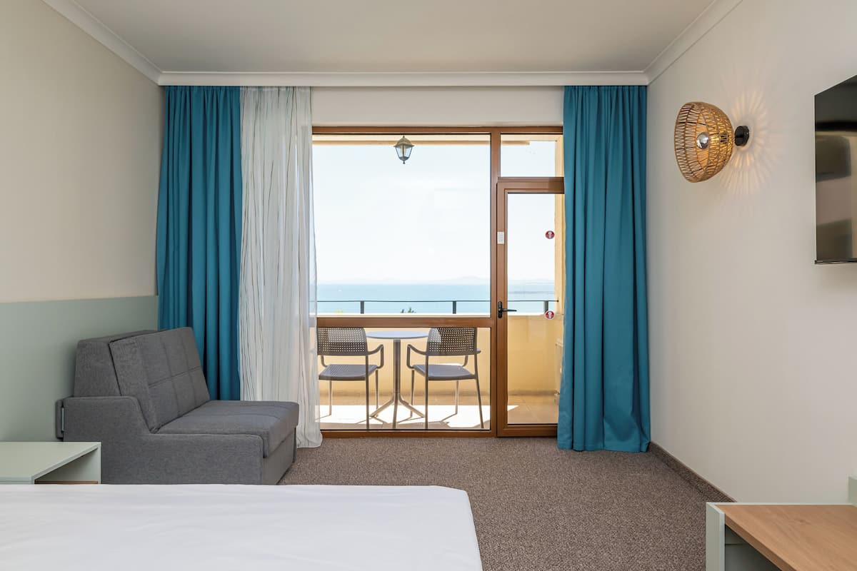 A hotel room with a bed, a sofa, a table, a TV, and a balcony with chairs and curtains.