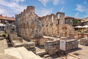 Ancient stone ruins with a signboard, surrounded by modern buildings and outdoor seating areas.