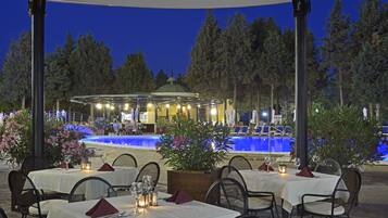 An outdoor dining area with tables set for two, surrounded by chairs, and a pool in the background.