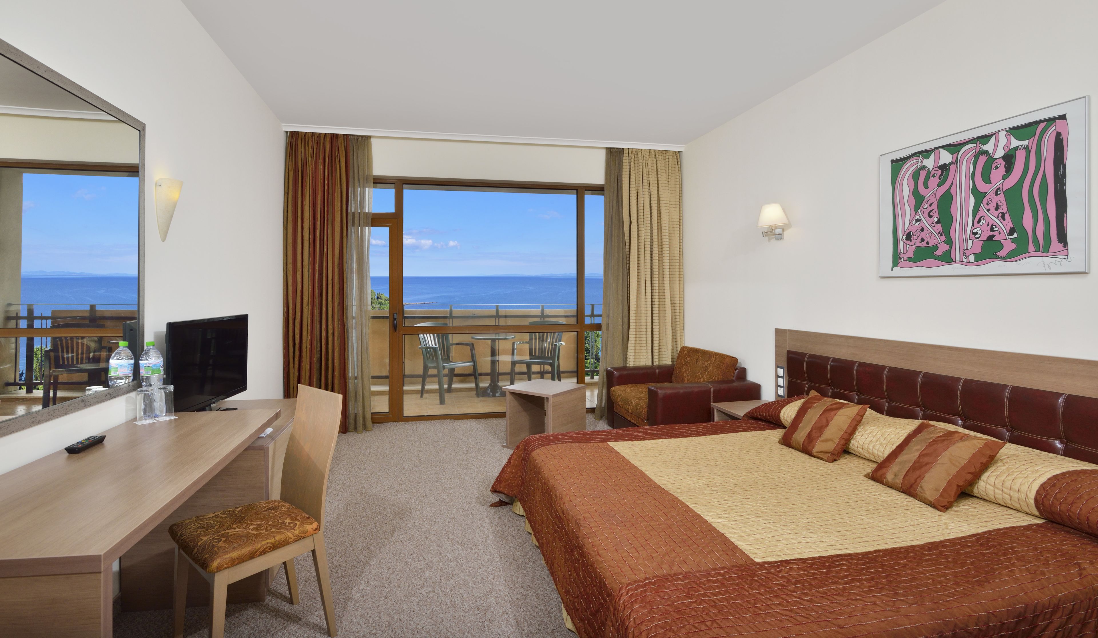 A hotel room with a bed, a desk, a chair, a television, and a balcony with a view of the sea.