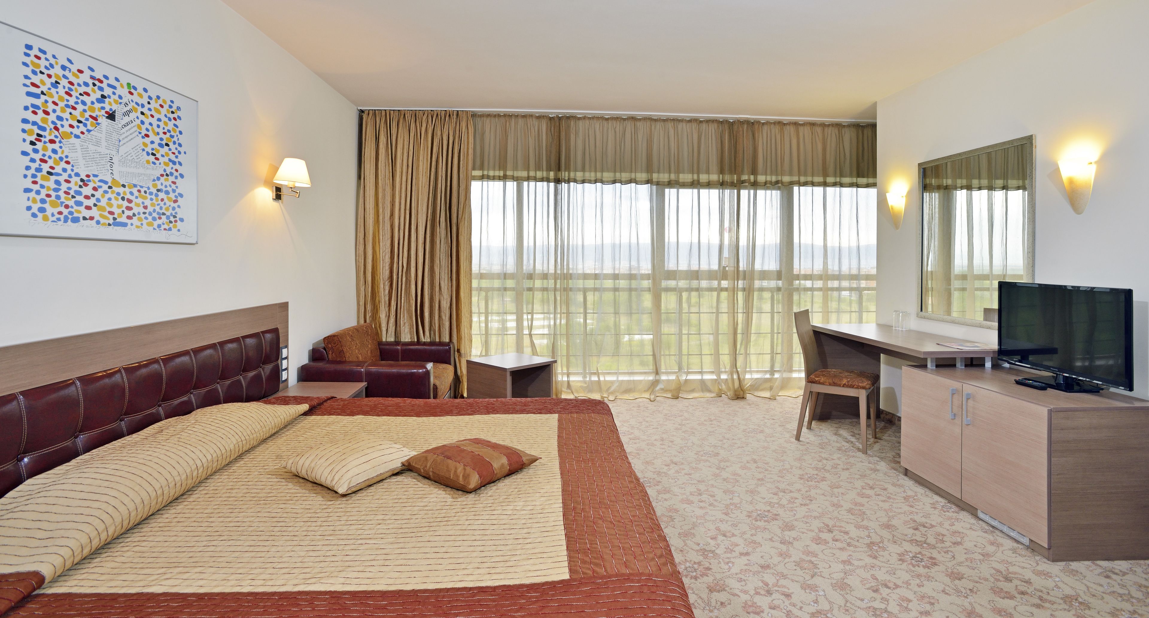 A hotel room with a large bed, a desk with a TV, a chair, a sofa, and a view of the outdoors.