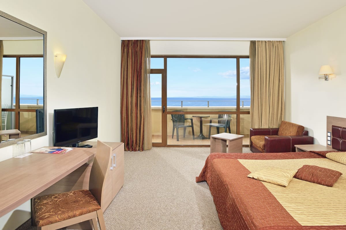 A hotel room with a large bed, a desk with a TV, a chair, a small table, and a balcony with a view of the sea.