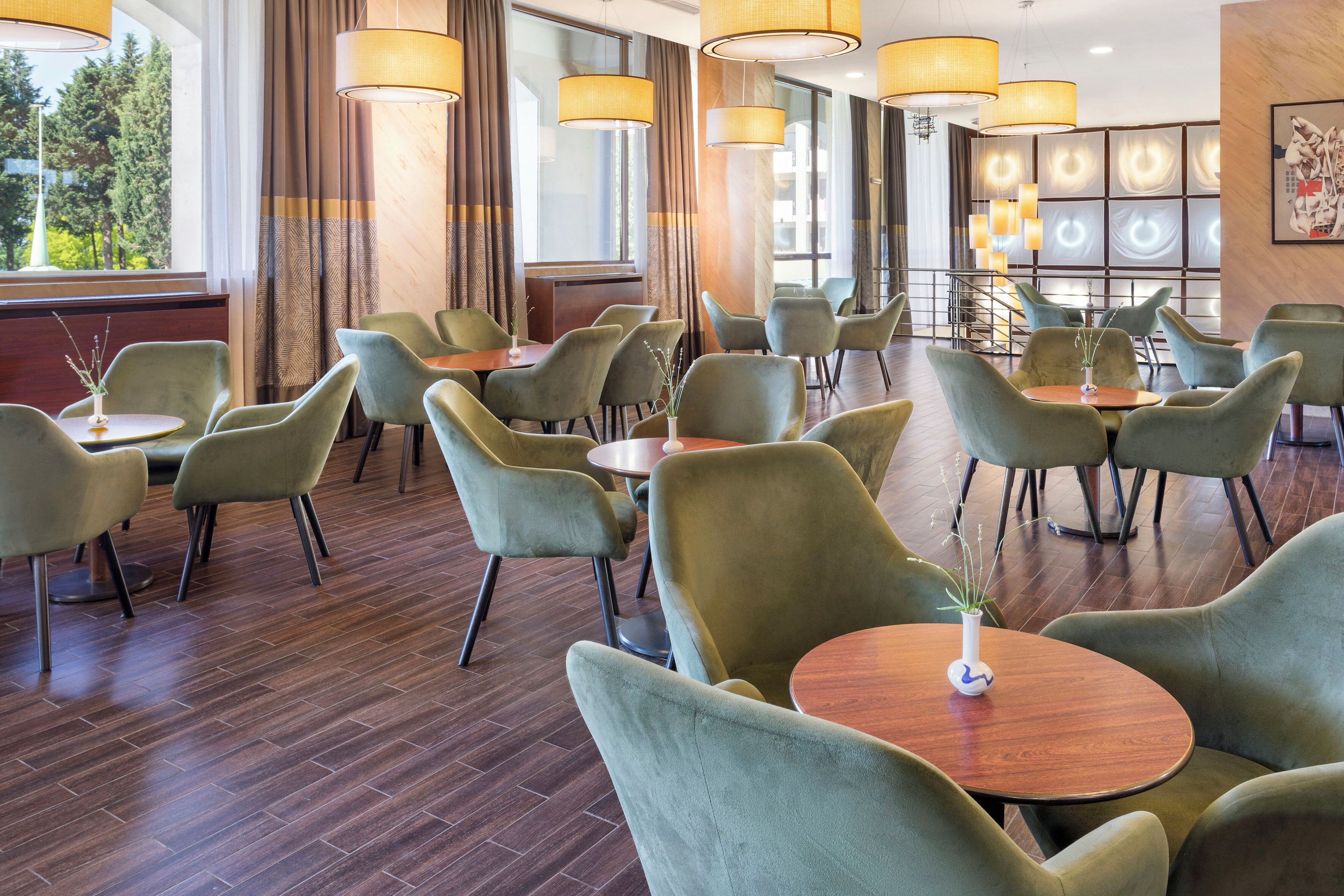 Un vestíbulo de hotel con sillones tapizados en verde, mesas redondas de madera y grandes lámparas colgantes.