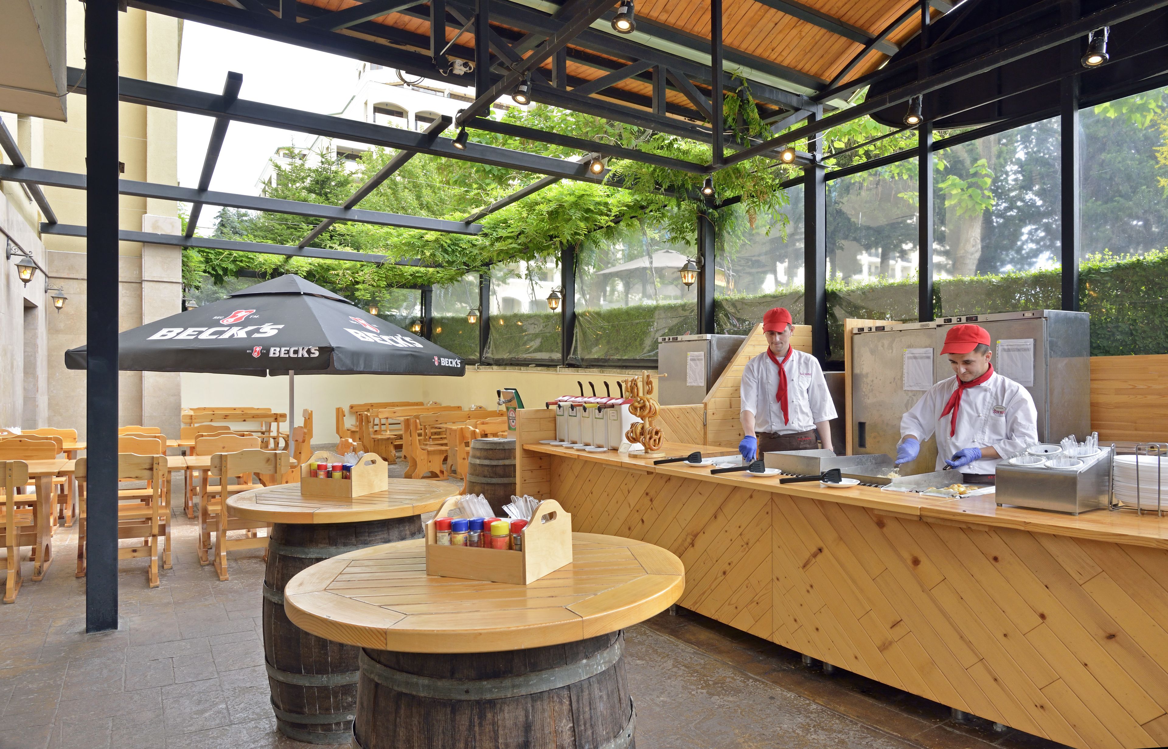 An outdoor café with wooden tables and chairs, a counter with a Beck's branded umbrella, and a large barrel.