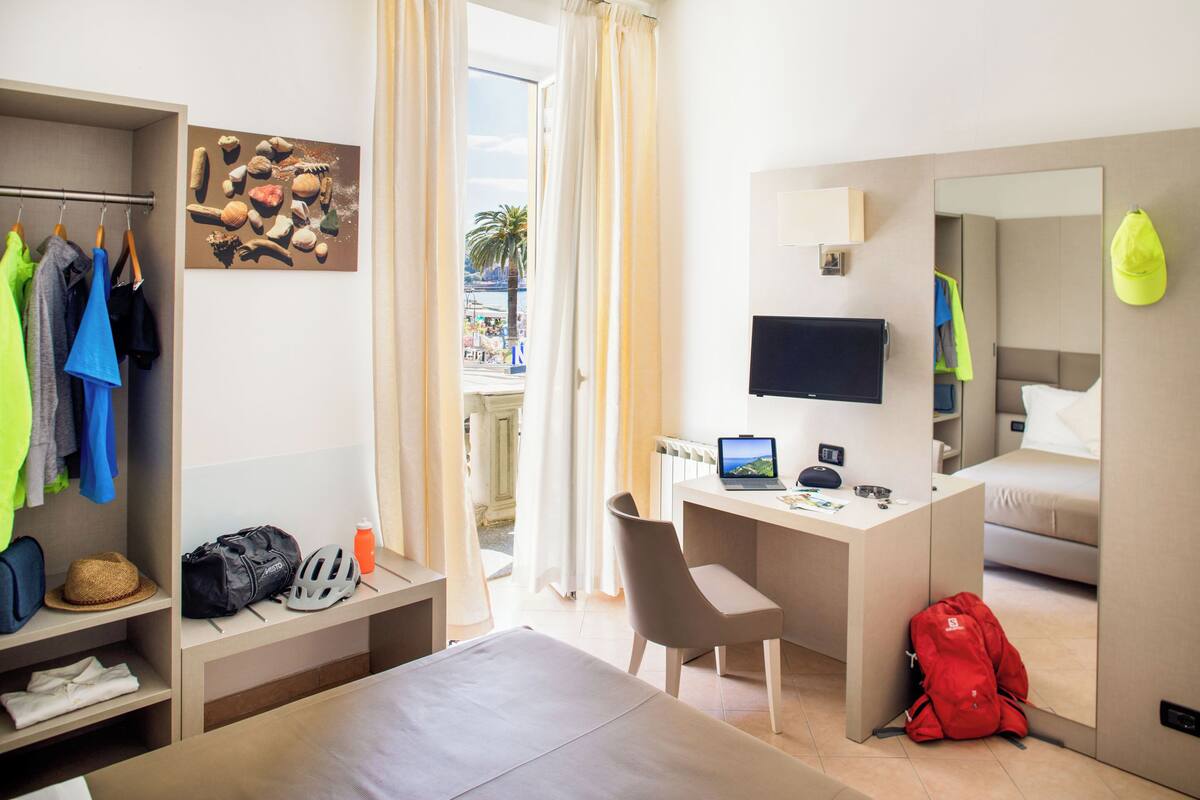 A hotel room with a bed, a desk with a laptop, a TV, a mirror, a wardrobe, and a window with curtains.
