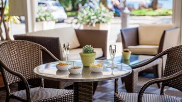 Une table ronde entourée de chaises en osier, une plante en pot et deux verres contenant un liquide transparent, probablement de l’eau ou du jus, sur une terrasse ensoleillée avec des sièges d’extérieur et une vue sur la rue.
