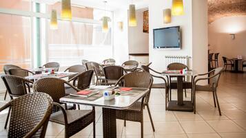 A dining area with wicker chairs and tables, a flat-screen TV on the wall, and large windows with blinds.
