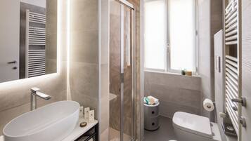 Un baño moderno con un lavamanos blanco de diseño, ducha con mampara de vidrio, inodoro y ventana con persianas.