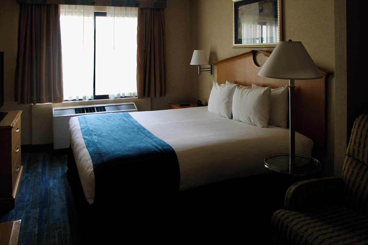 A hotel room with a large bed, a bedside table, a lamp, a chair, and a window with curtains.