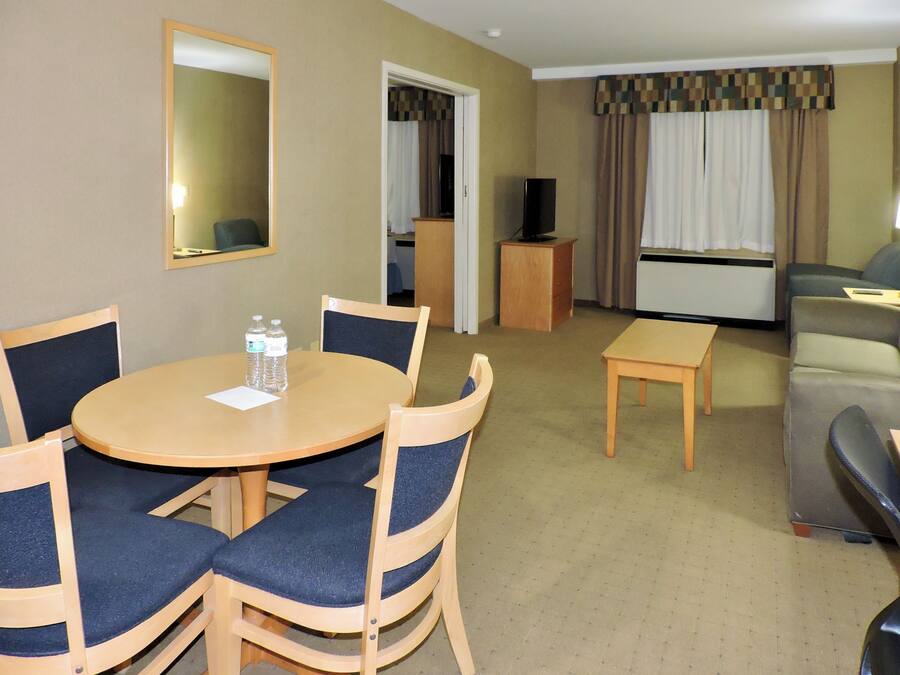 Habitación de hotel con una mesa redonda de comedor, cuatro sillas, un sofá, una mesita, un televisor sobre un mueble y una ventana con cortinas.