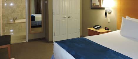 A hotel room with a bed, a bedside table with a phone and alarm clock, a mirror, a door, and a bathroom with a sink and mirror.