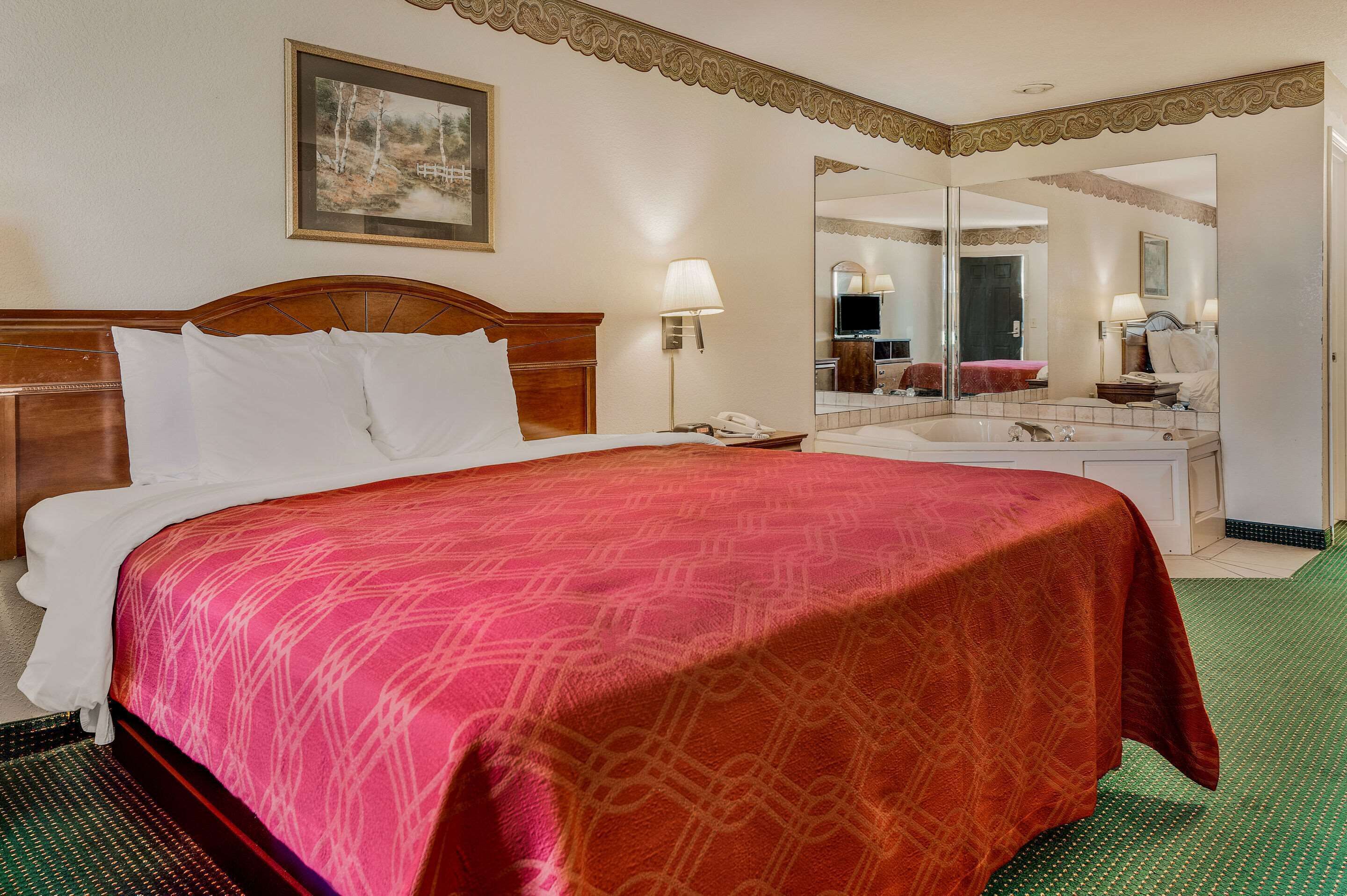 Quarto de hotel com uma cama grande, uma mesa de cabeceira com abajur, um espelho e um banheiro visível por uma porta aberta.