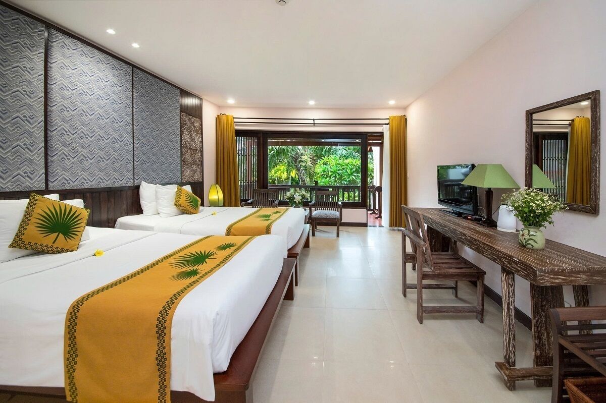 Quarto de hotel com duas camas, uma mesa de jantar, televisão e varanda com vista para vegetação.