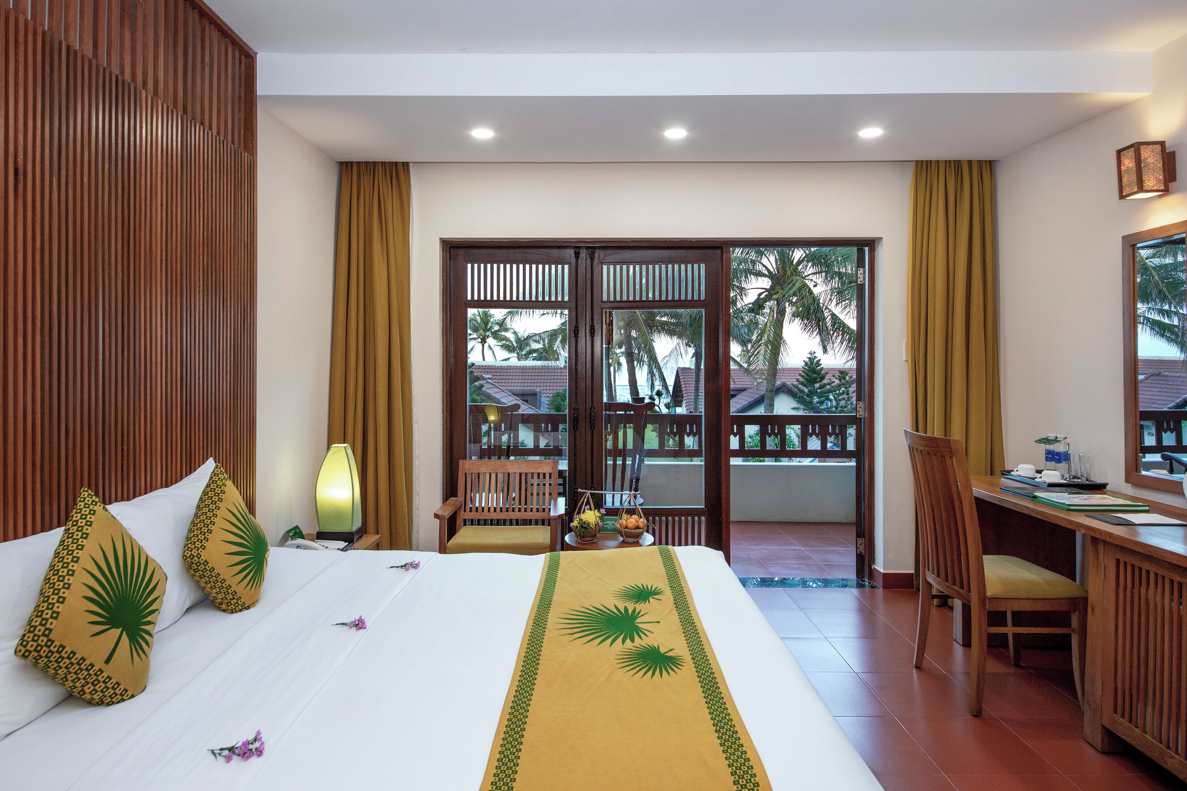 A hotel room with a bed, a desk, a chair, a balcony with a view of palm trees, and a wooden partition.