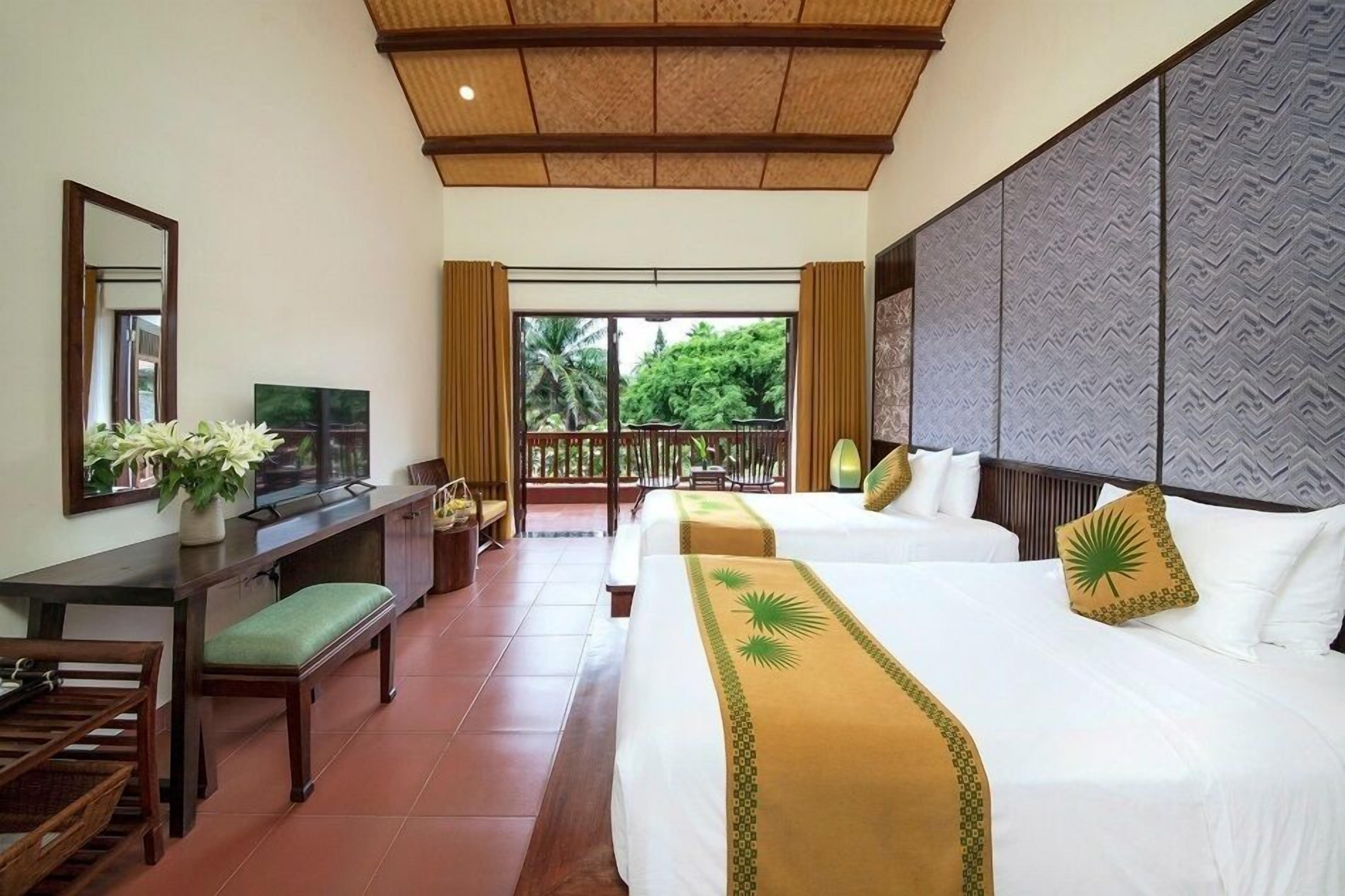 A hotel room with two beds, a desk with a chair, a television, and a balcony with a view of greenery.