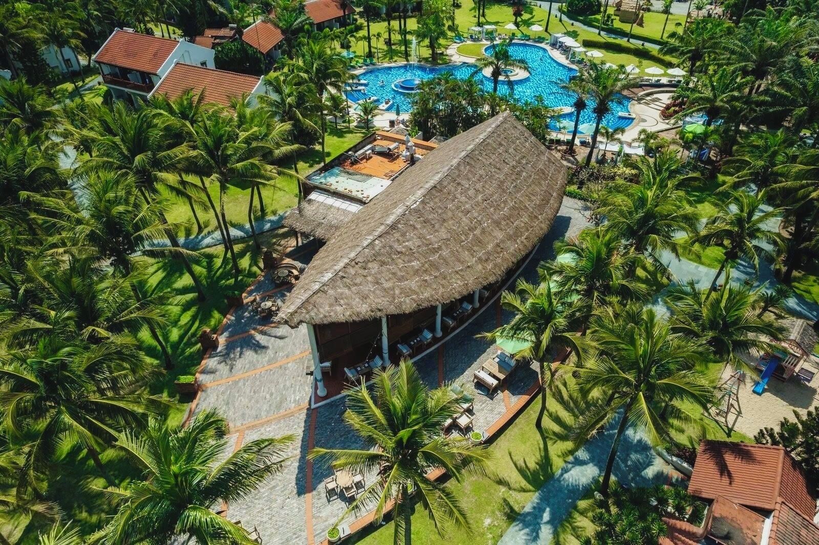 Un complexe hôtelier doté d’une grande piscine, de palmiers et d’un bâtiment à toit de chaume.