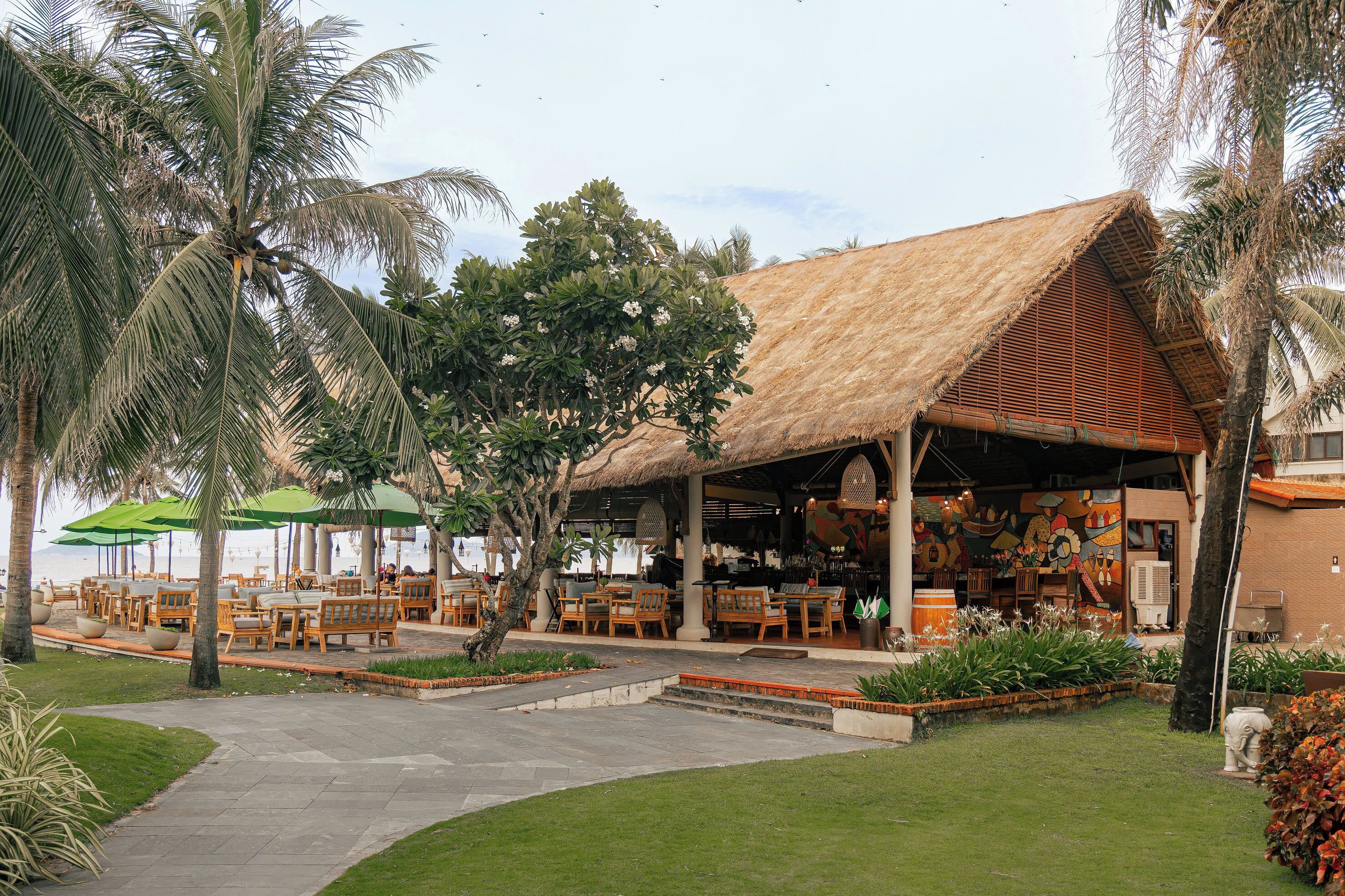 An outdoor seating area with wooden tables and chairs, surrounded by palm trees and a thatched-roof structure.