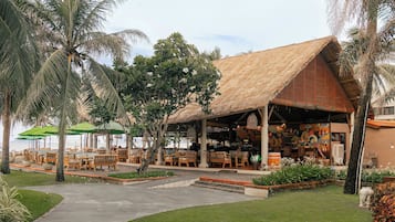 An outdoor seating area with wooden tables and chairs, surrounded by palm trees and a thatched-roof structure.
