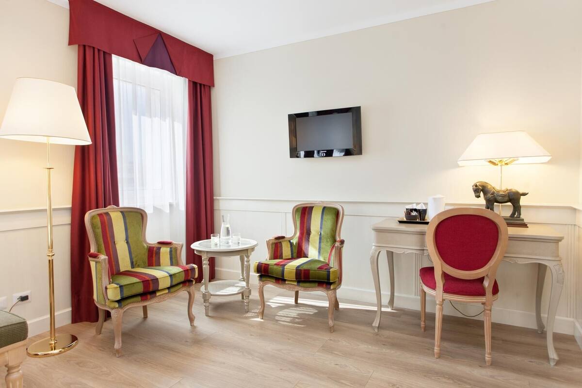 A hotel room with two striped armchairs, a small round table, a desk with a lamp, and a mounted TV.