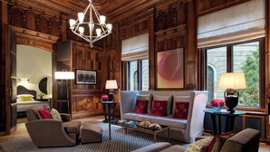 A spacious living room with a large sofa, armchairs, and a coffee table. There is a chandelier, a fireplace, and a large window with a view. - Hotel De Rome Berlin (Berlin)