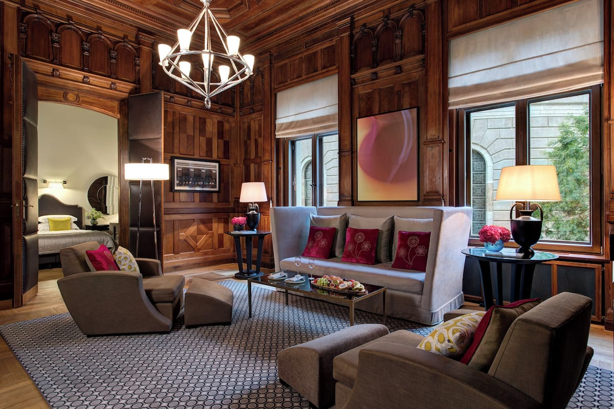 A spacious living room with a large sofa, armchairs, and a coffee table. There is a chandelier, a fireplace, and a large window with a view.