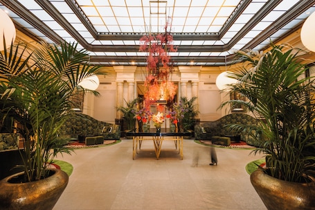 A spacious lobby with a central table, potted plants, and a high ceiling with a skylight.. Hotel De Rome Berlin