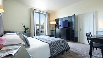Habitación de hotel con cama, escritorio con silla, televisor, una pequeña mesa con planta y vistas al balcón exterior.