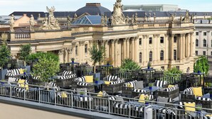 A rooftop terrace with outdoor seating and a view of a historic building with statues. - Hotel De Rome Berlin (Berlin)