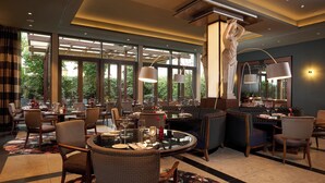 A well-lit dining area with round tables, comfortable seating, and large windows offering a view of the outdoors. - Hotel De Rome Berlin (Berlin)