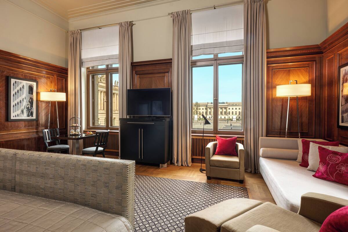A hotel room with a large window, a sofa, a TV, and a dining area.