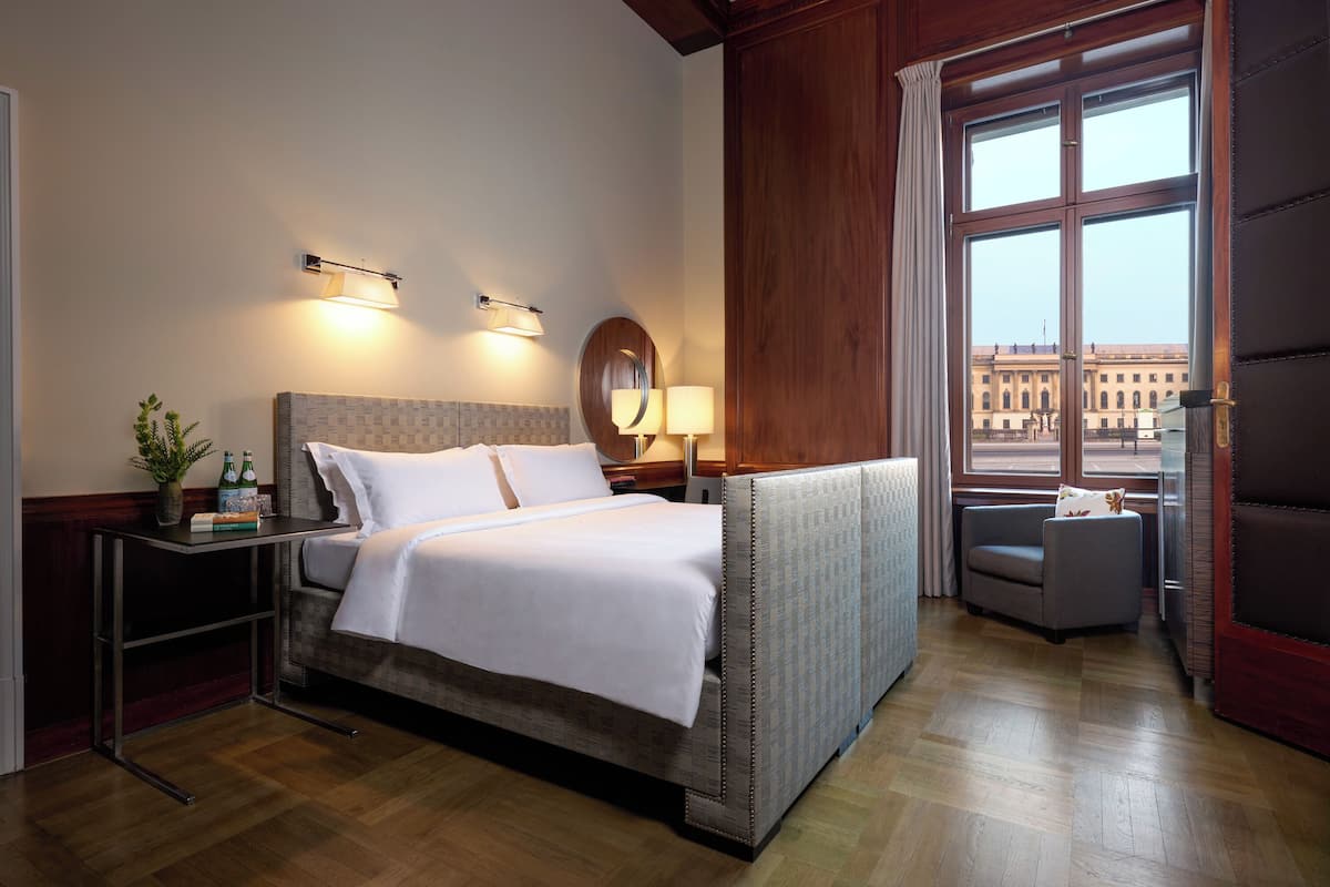 A hotel room with a bed, bedside tables, a lamp, a view of a building through a window, and a chair.