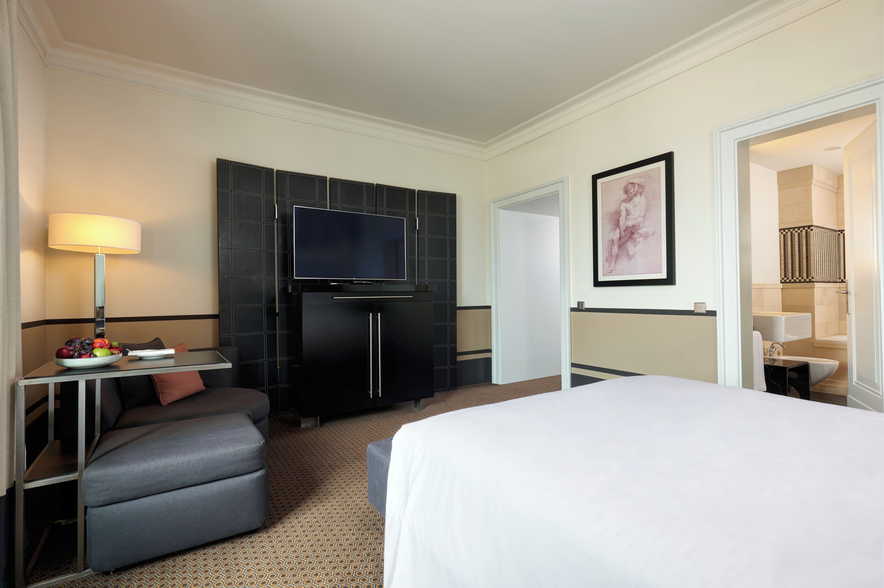 A hotel room with a bed, a desk with a lamp, a television, and a framed artwork on the wall.