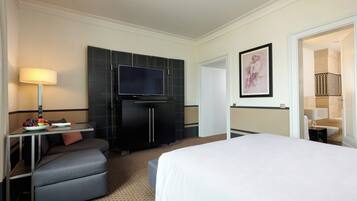 A hotel room with a bed, a desk with a lamp, a television, and a framed artwork on the wall.
