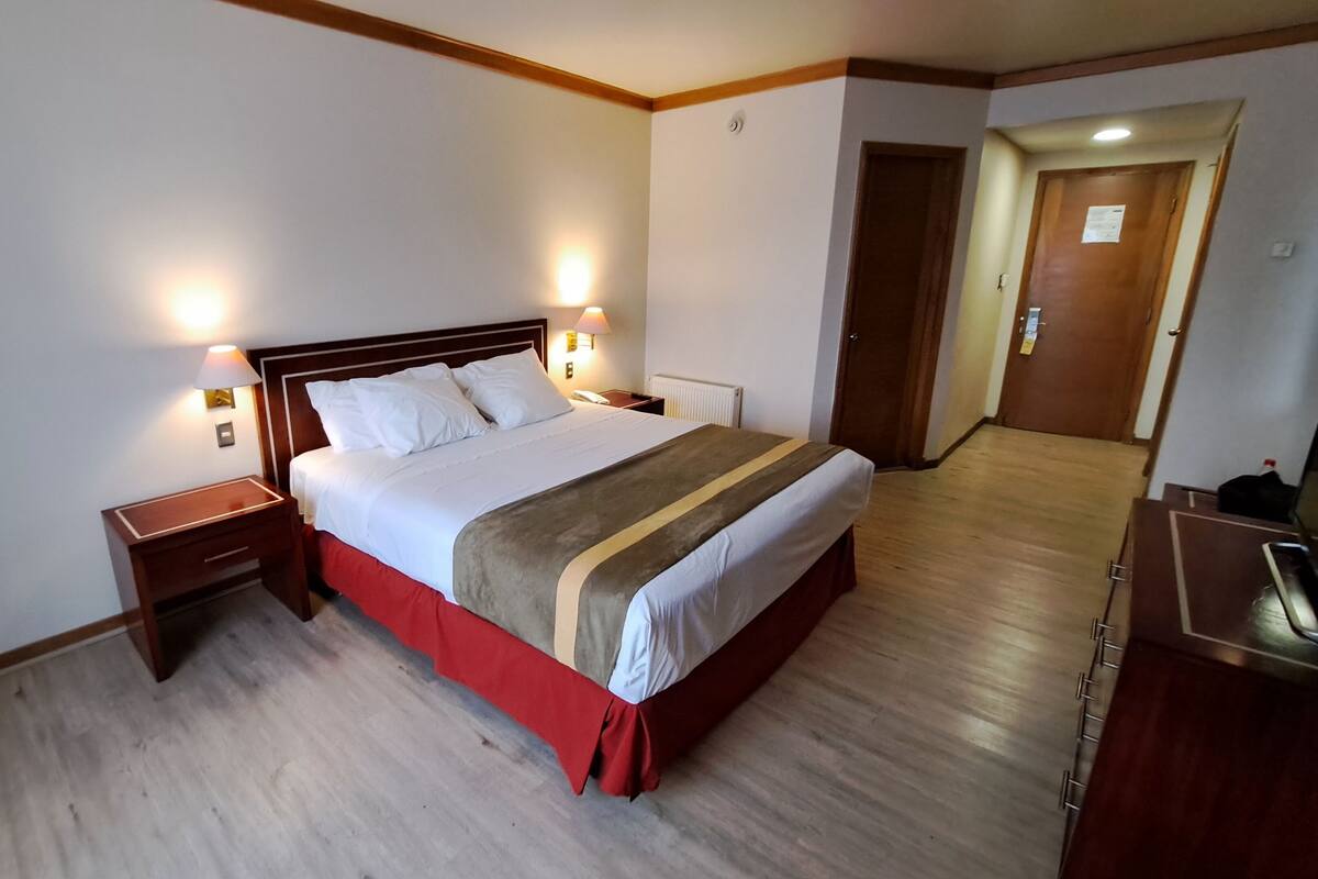 A hotel room with a large bed, two bedside lamps, a wooden nightstand, and a red bed runner.