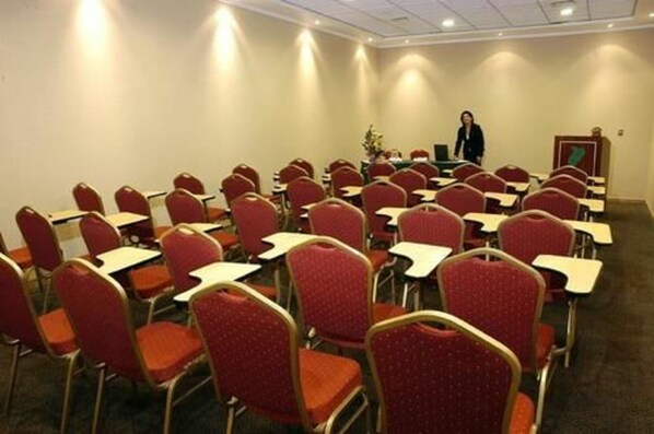 Un salón de conferencias con filas de sillas rojas y mesas, un atril, y una persona de pie al frente.