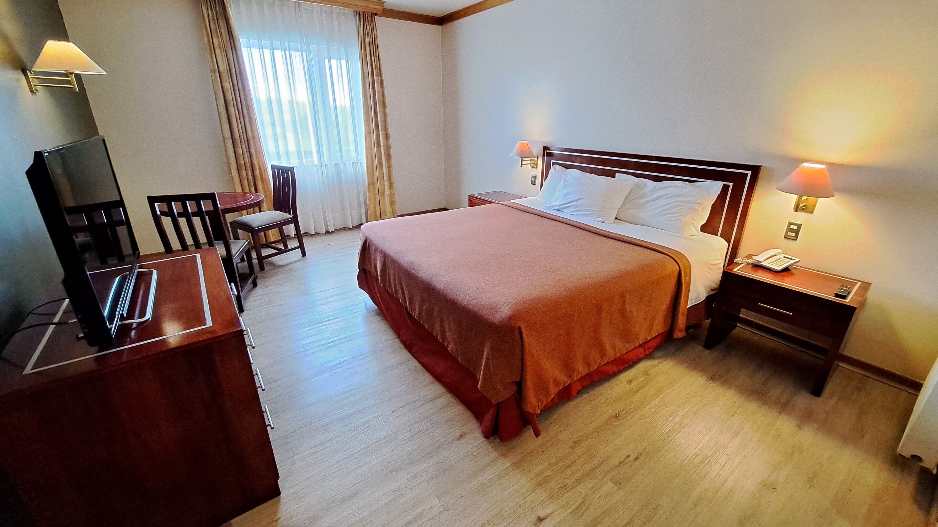 Una habitación de hotel con cama, mesitas de noche, un escritorio con computadora, una silla y una ventana con cortinas.