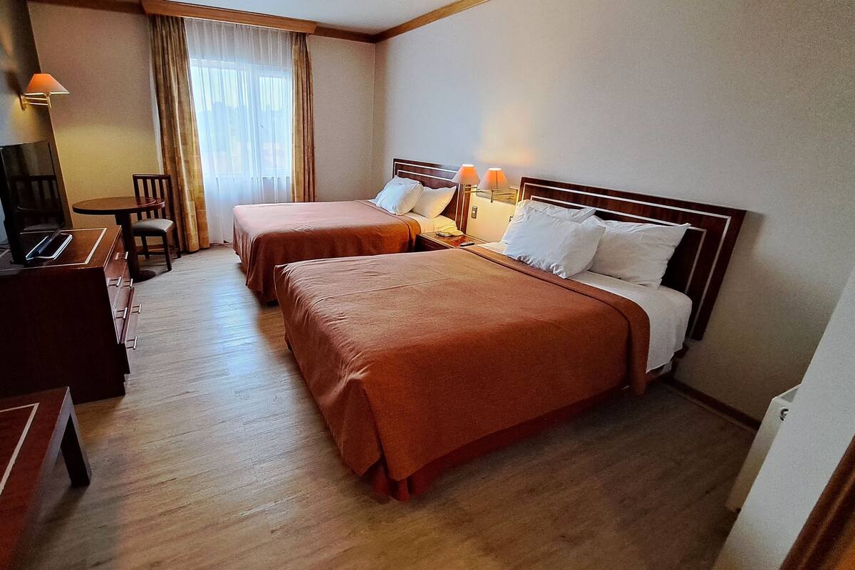 A hotel room with two beds, a desk with a chair, a lamp, and a window with curtains.