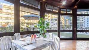 Uma área de refeições com uma mesa posta para dois, rodeada por cadeiras brancas, um vaso de planta e vista para um conjunto de edifícios através de grandes janelas.