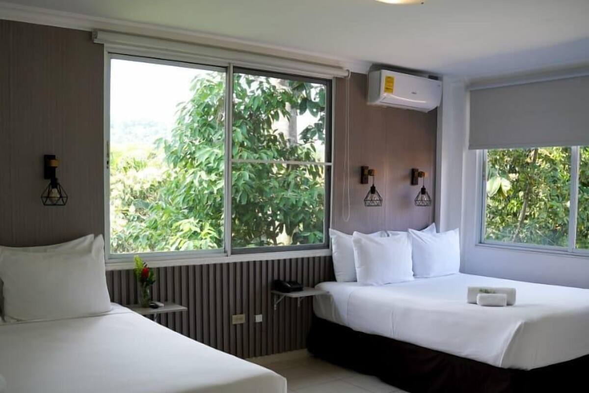 A hotel room with two beds, a large window, a radiator, and a ceiling light.
