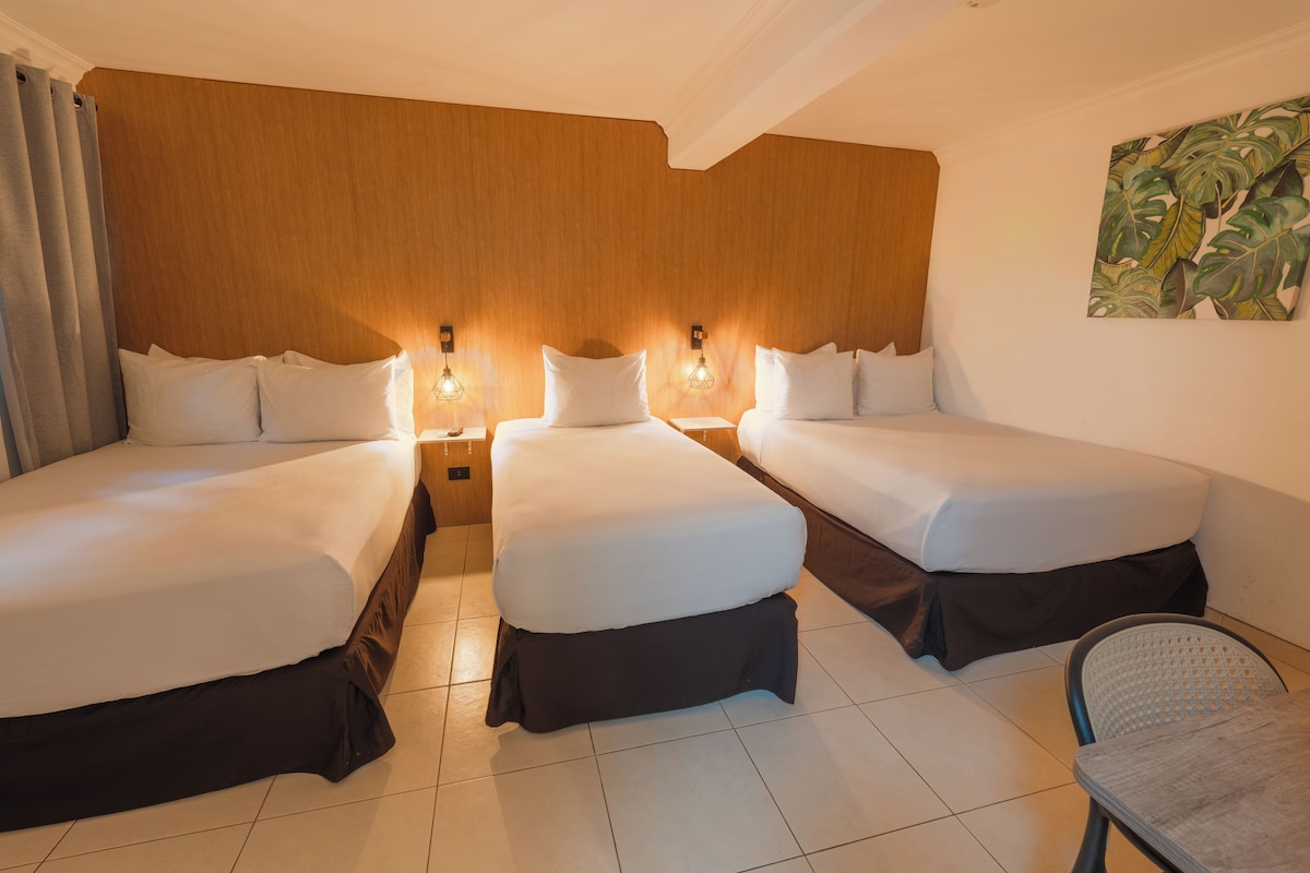 Quarto de hotel com três camas de solteiro, cabeceira de madeira, duas luminárias de mesa, uma cadeira e um quadro na parede.