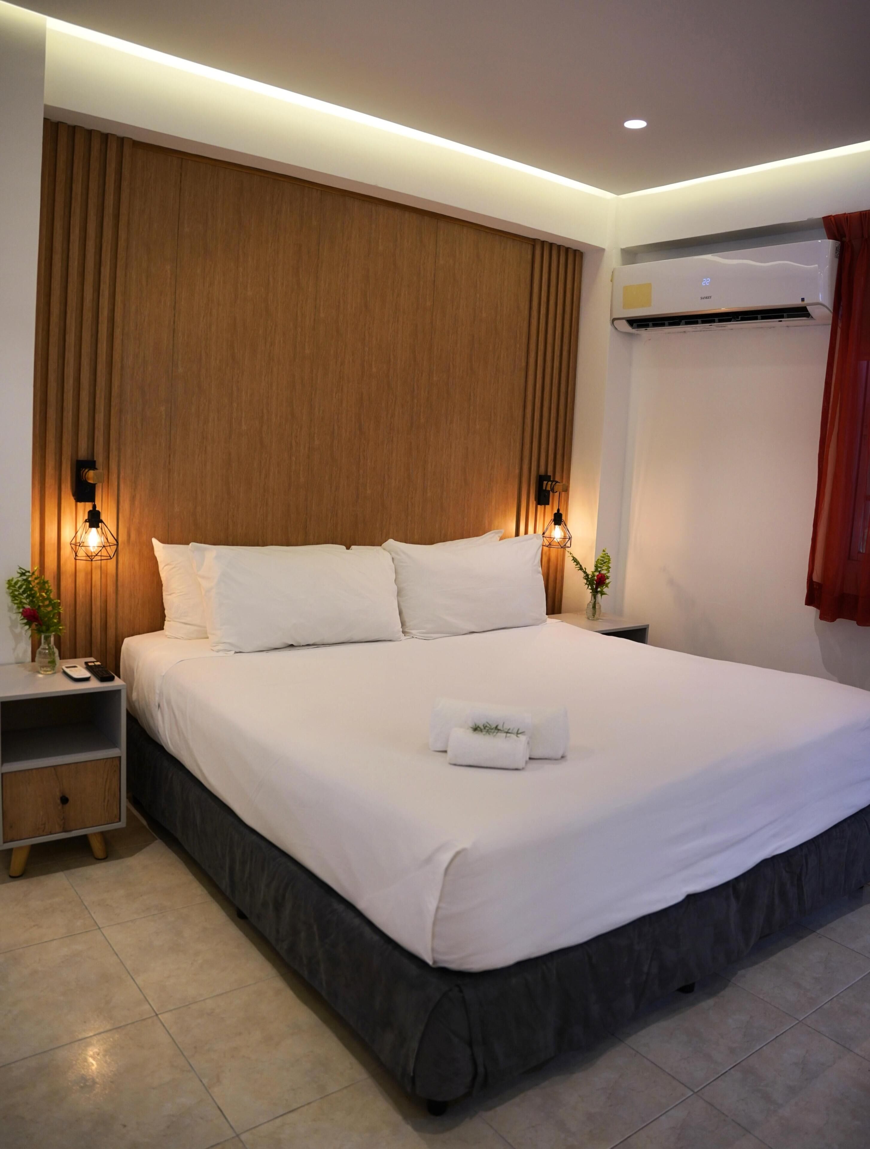 A modern hotel room with a large bed, wooden headboard, bedside lamps, a nightstand, and an air conditioning unit.